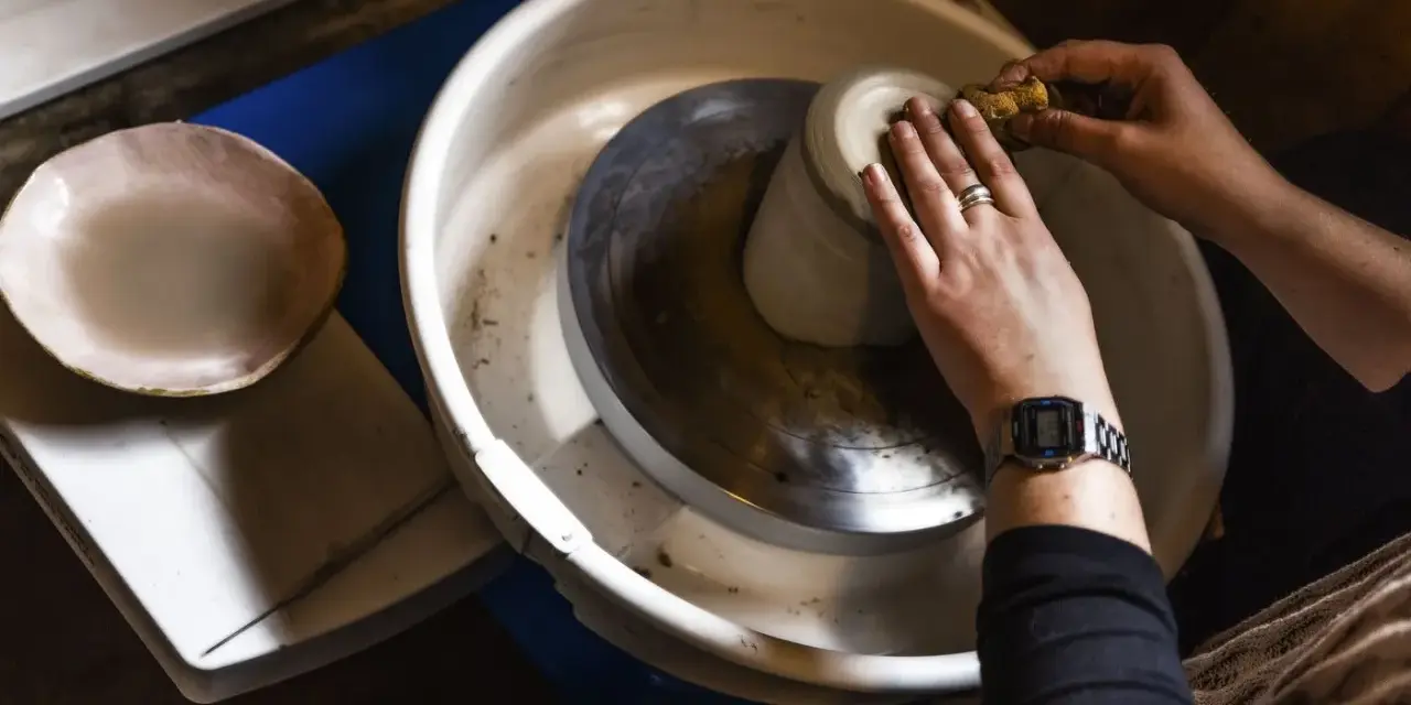 Händer som formar en mugg på en drejskiva. En svamp används för att jämna till leran medan man drejar lera.