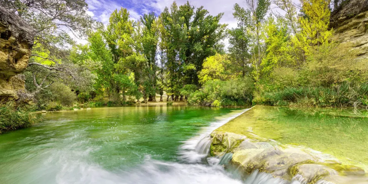 La poza de doña urraca, un remanso de agua verde esmeralda con una pequeña cascada, rodeada de frondosa vegetación y árboles altos bajo un cielo azul.