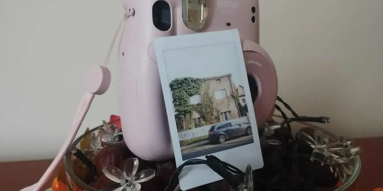 Różowy aparat Instax z wydrukowanym zdjęciem domu i samochodu. Idealny pomysł, jak zrobić prezent ze zdjęć, ozdobiony jesiennymi liśćmi i lampkami.