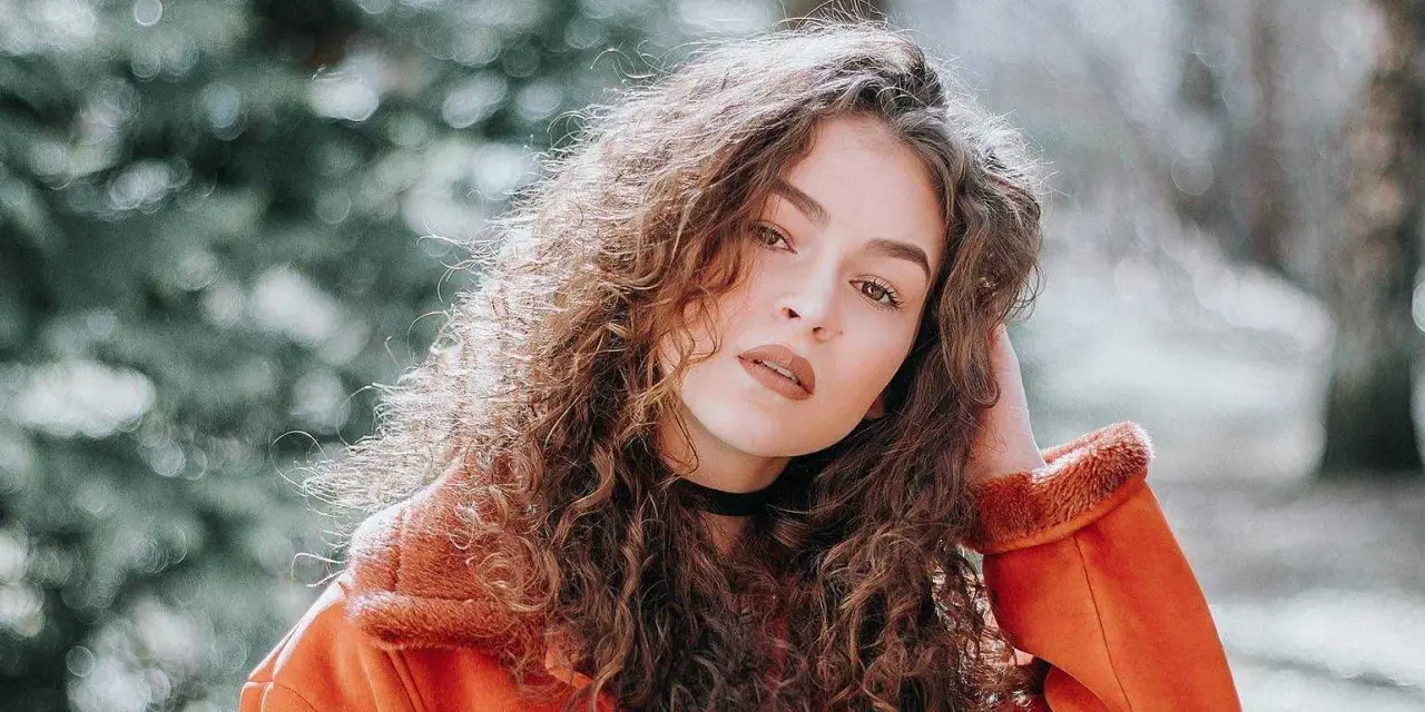 Jeune femme aux cheveux bouclés, portant une veste orange. La retouche photo netteté en ligne sublime son regard intense.