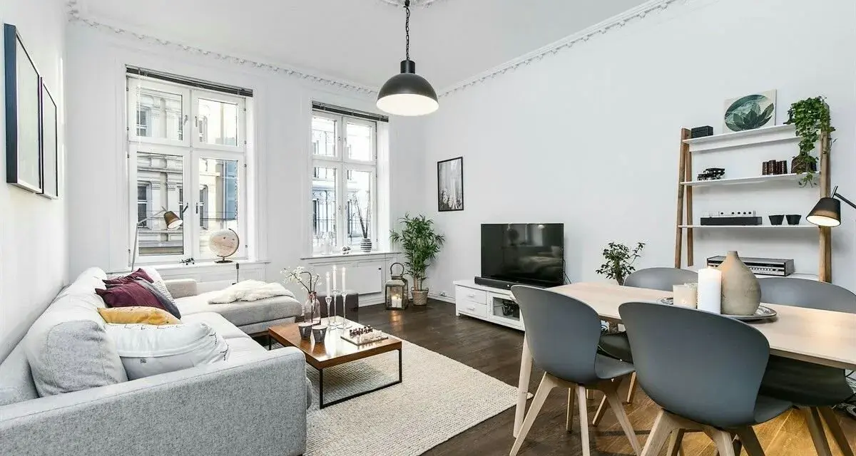 Elegante salón con sofá gris, mesa de centro y comedor. El **suelo oscuro** de madera contrasta con las paredes blancas y la luz natural.