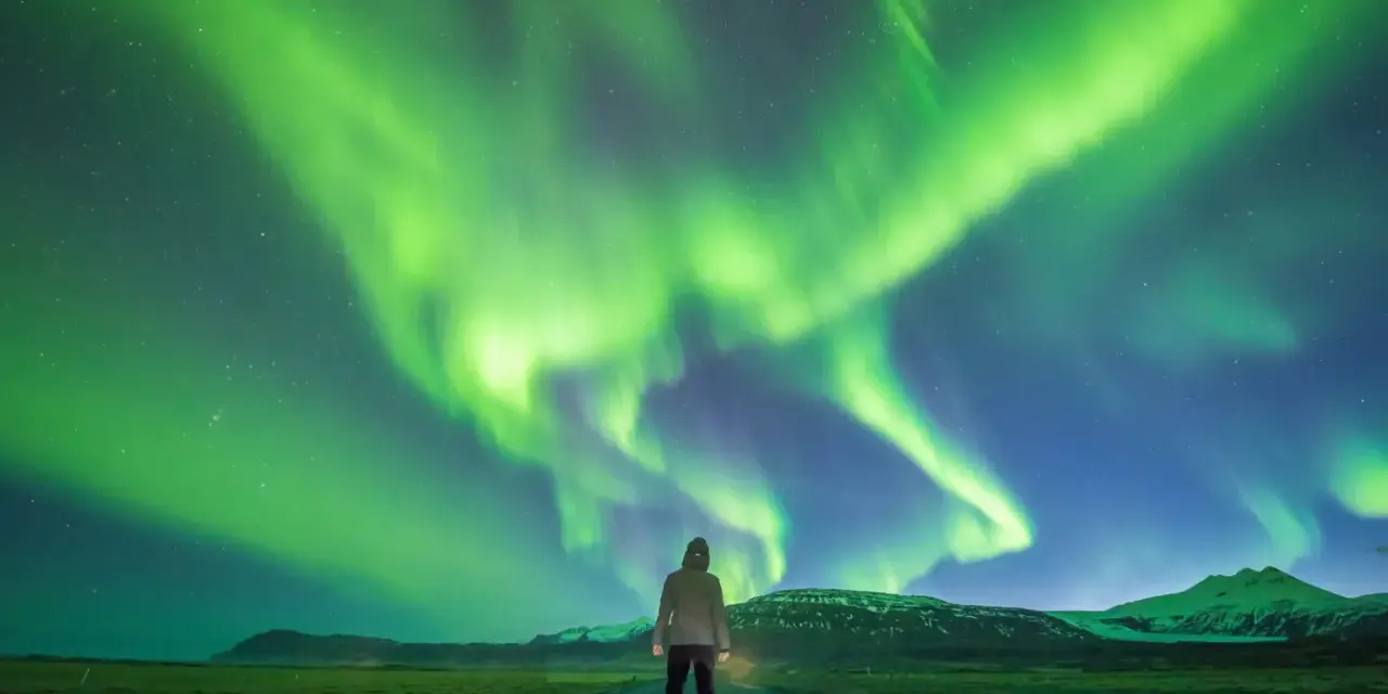Człowiek stoi na drodze pod rozgwieżdżonym niebem, gdzie tańczy zorza polarna. Islandia, październik, pogoda idealna na takie widowisko.