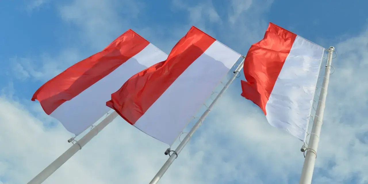 Trzy powiewające na wietrze flagi, każda z czerwono-białą szarfą, na tle błękitnego nieba z chmurami.