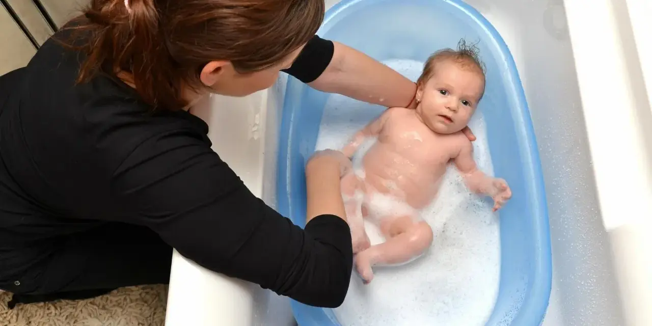 Mama delikatnie myje noworodka w niebieskiej wanience. Maluch pluska się w pianie, a mama uśmiecha się do niego.