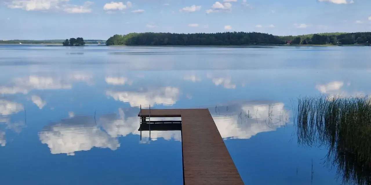 Drewniane molo na spokojnym jeziorze Wysockim, odbijającym błękitne niebo z białymi chmurami.