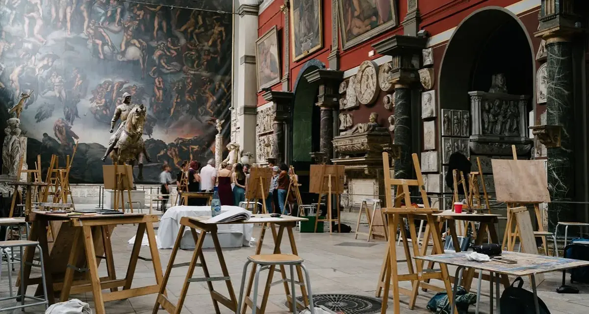 Des chevalets et des toiles dans une **école d'art appliqués**, devant une fresque monumentale et des sculptures. Des étudiants observent une œuvre.