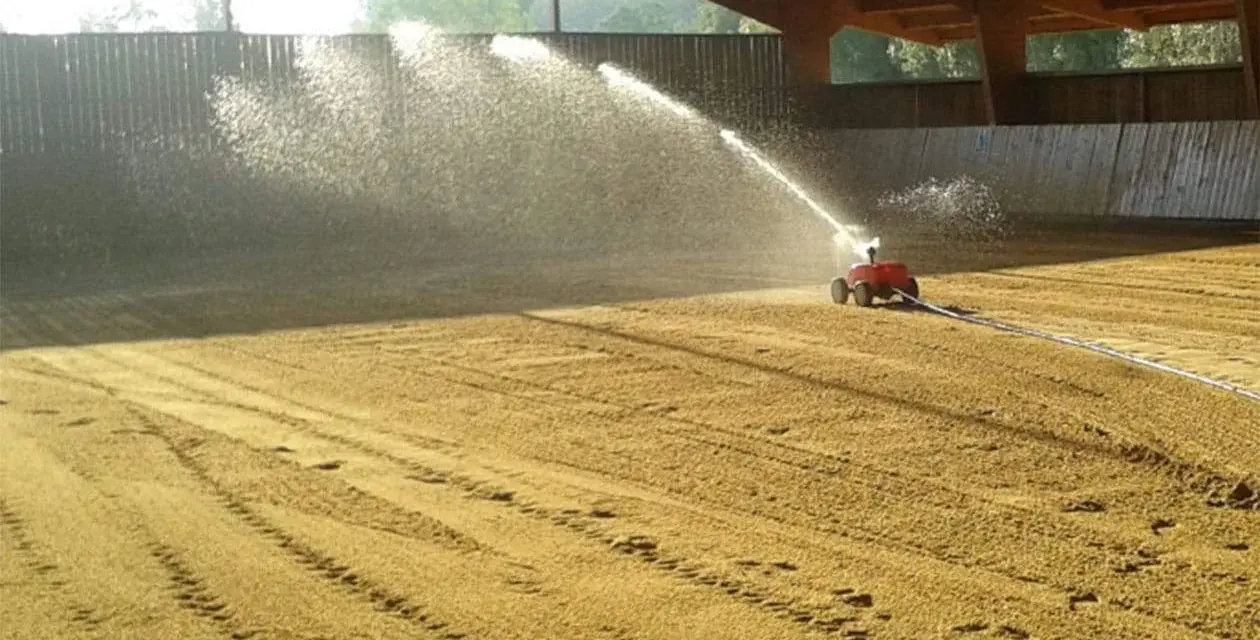 Arrosage d'une carrière équestre par un système mobile, créant un voile d'eau sur le sable.