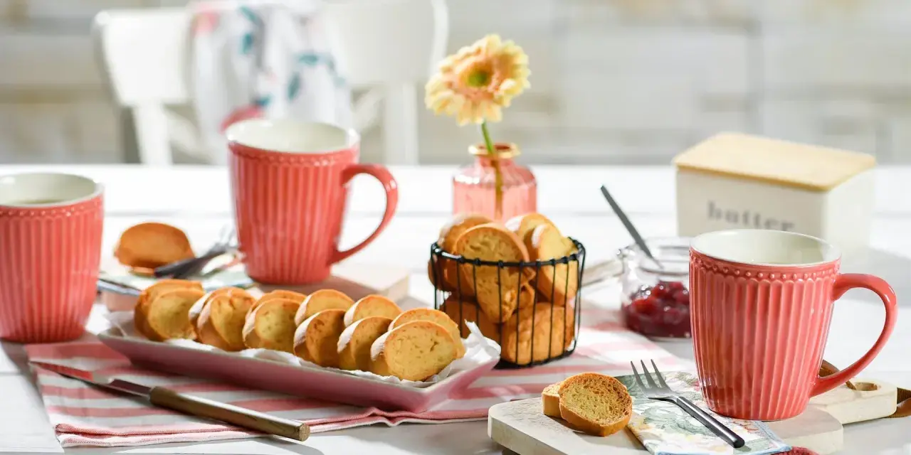 Leckeres Zwieback-Rezept: goldbraune Scheiben auf rosa Platte, dazu Marmelade, Butterdose und rote Tassen.