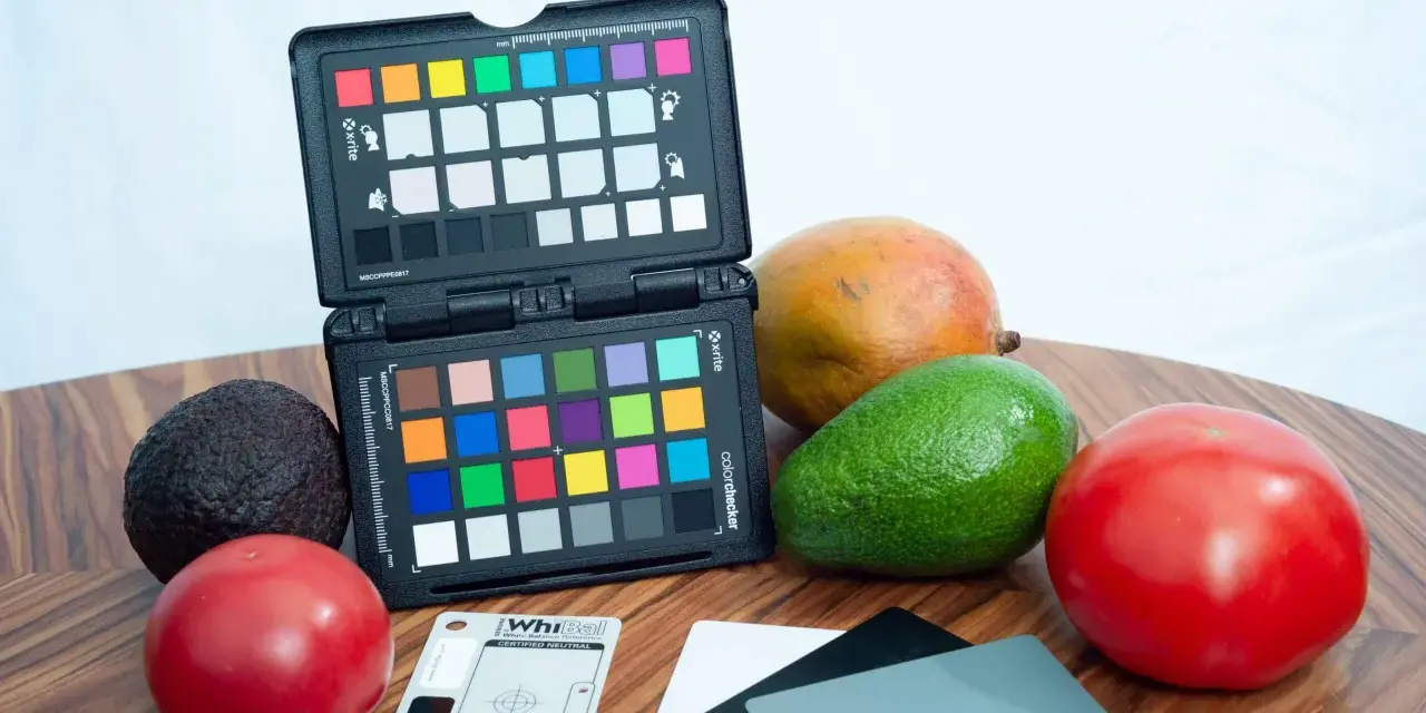 Na drewnianym blacie leżą owoce: awokado, mango, pomidory. Obok znajduje się otwarty colorchecker z kolorowymi polami i szara karta do kalibracji balansu bieli.