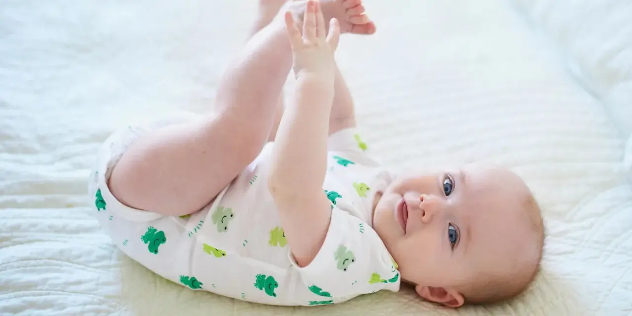 Ein süßes Baby, ca. 6 Monate alt, liegt auf dem Rücken und spielt mit seinen Füßen. Es trägt einen weißen Body mit grünen Fröschen.