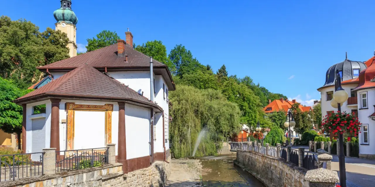 Urokliwy strumyk w Polanicy Zdrój, otoczony kamiennymi murkami i zielenią. W tle widać wieżę kościelną i kolorowe kamieniczki.