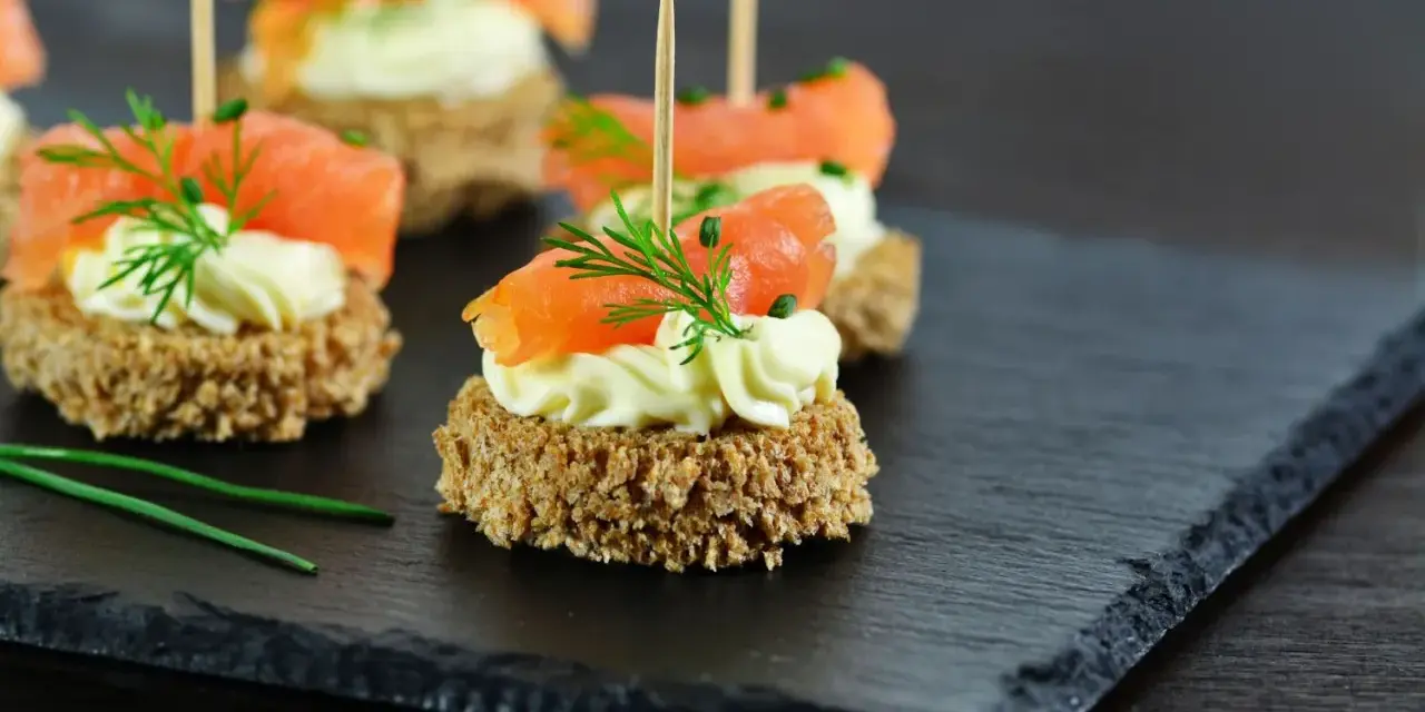 Eleganckie przystawki na komunię: krążki ciemnego pieczywa z kremowym serkiem, łososiem i koperkiem.