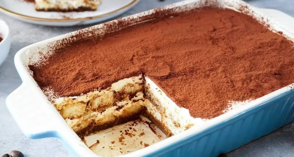 Ein köstliches Tiramisu rezept ohne alkohol, bestäubt mit Kakao, in einer blauen Auflaufform. Ein Stück ist bereits entnommen.