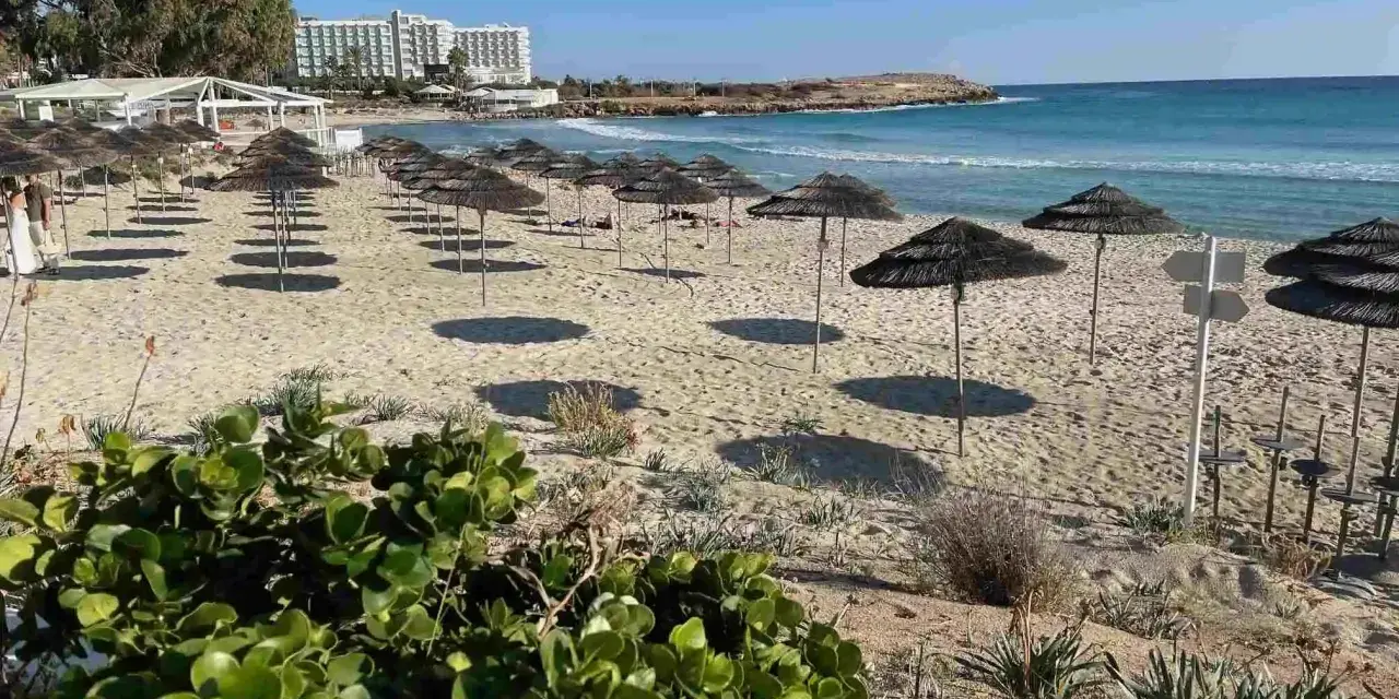 Puste cypr plaże z rzędami parasoli i hotel w tle. Słońce rzuca długie cienie na piasek.