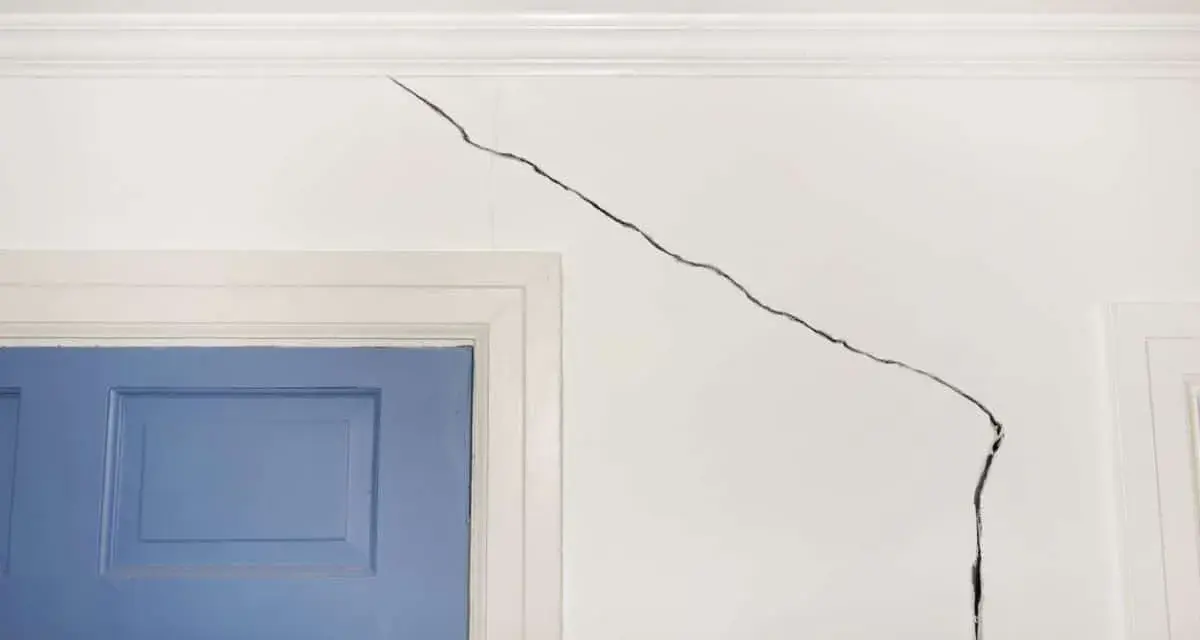 Grosse fissure sur mur intérieur blanc, près d'une porte bleue. Il faut réparer cette fissure pour un intérieur impeccable.