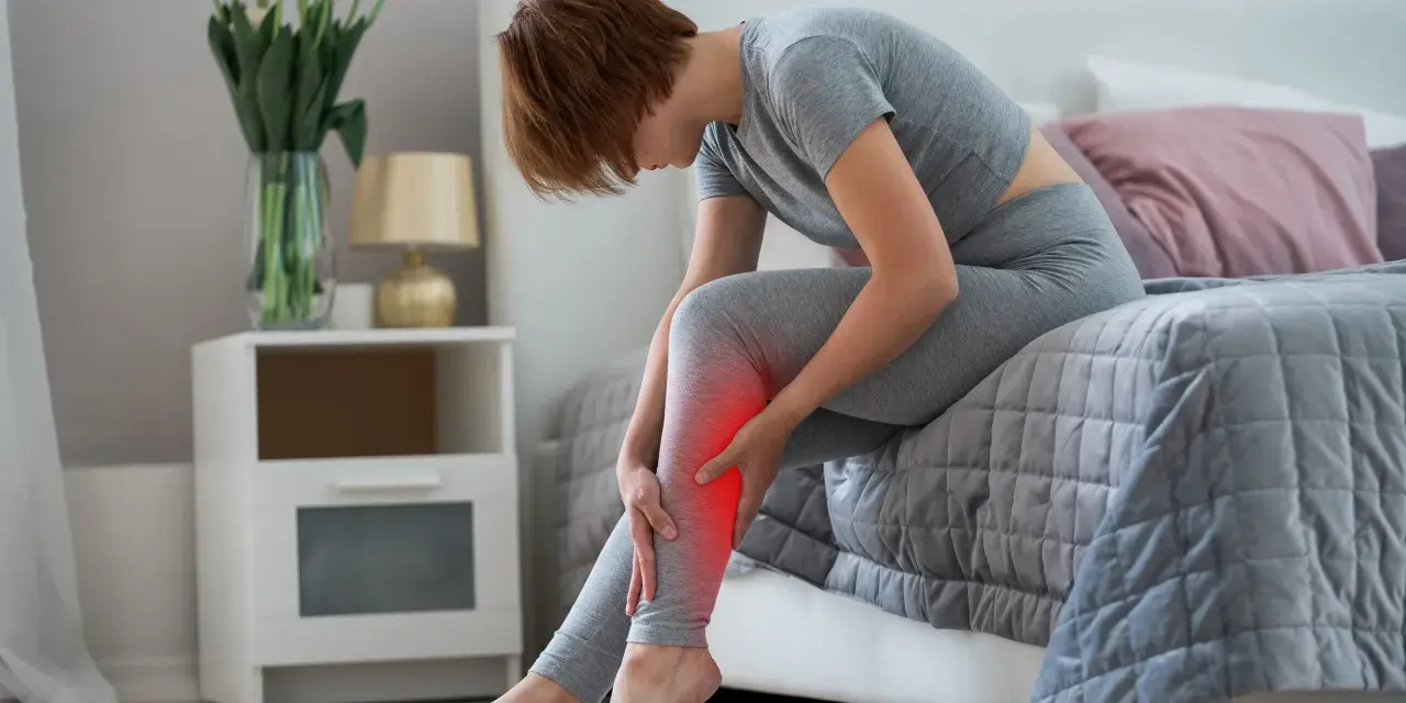 Femme assise sur un lit, se tenant la jambe douloureuse, souffrant de crampes nocturnes.