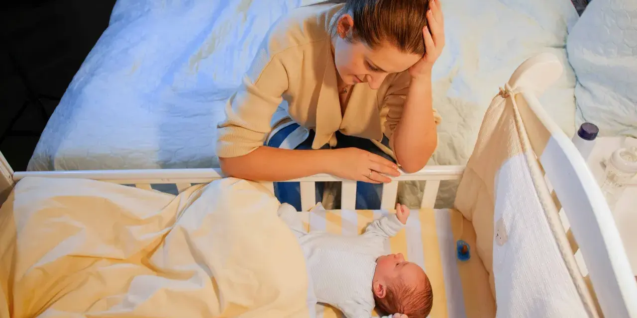 Zmęczona mama pochyla się nad łóżeczkiem, zastanawiając się, kiedy nastąpi regres snu. Niemowlę śpi spokojnie.