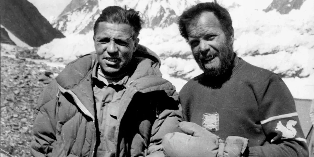 Zwei Bergsteiger, die die K2 Erstbesteigung schafften, lächeln in die Kamera. Im Hintergrund sind schneebedeckte Berge zu sehen.