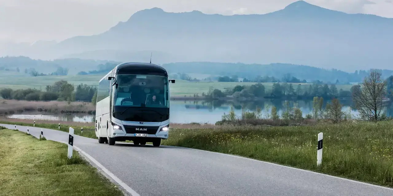 Biały **bus** marki **MAN** jedzie krętą drogą nad jeziorem, w tle góry.