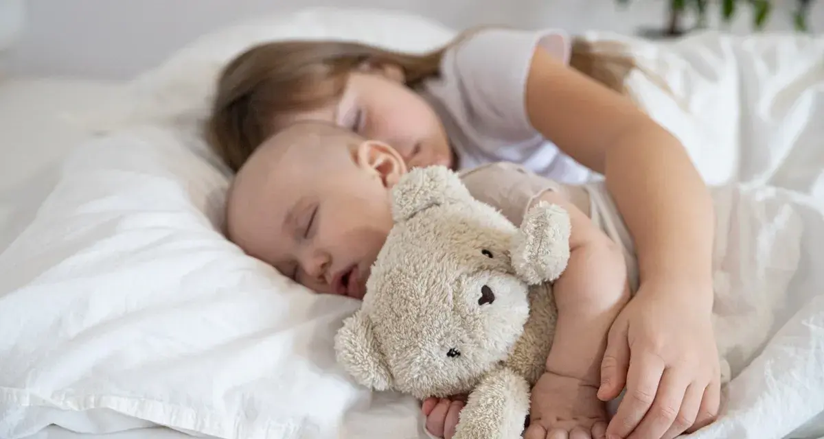 Dzieci śpią wtulone w misia. Starsza siostra czuwa nad maluszkiem, pokazując, ile śpi dwulatek i jak ważny jest sen dla rozwoju.