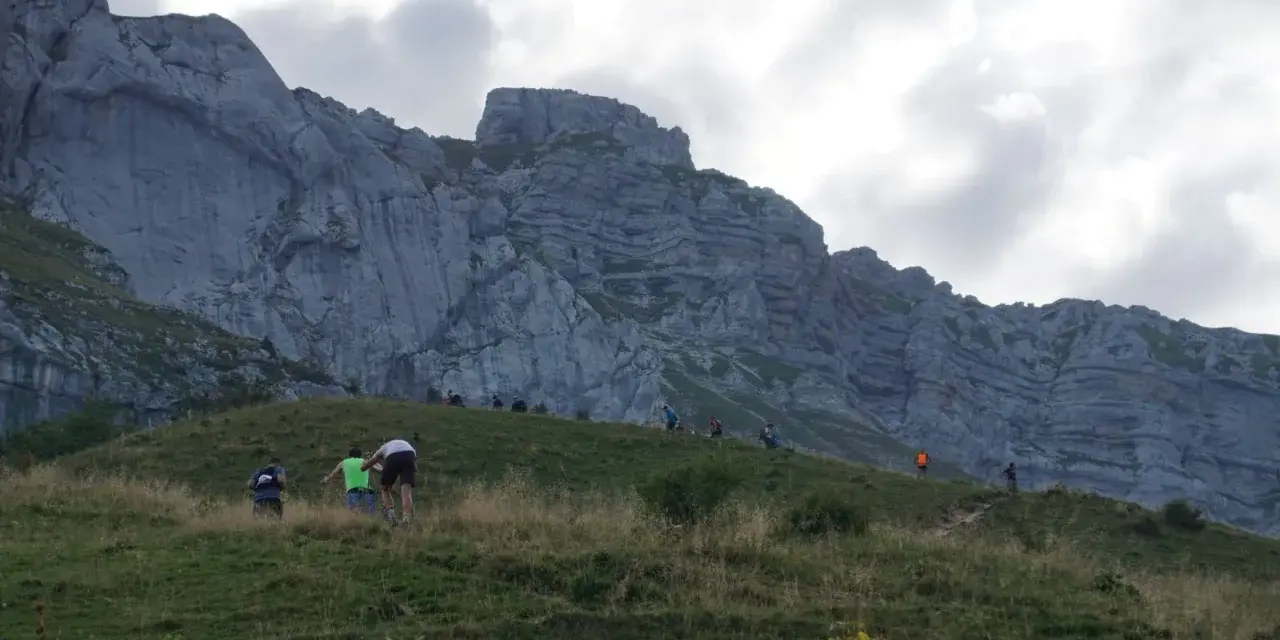 Randonneurs sur une pente herbeuse face à une falaise imposante. Le chemin sinueux et la hauteur suggèrent que la tournette est dangereuse.