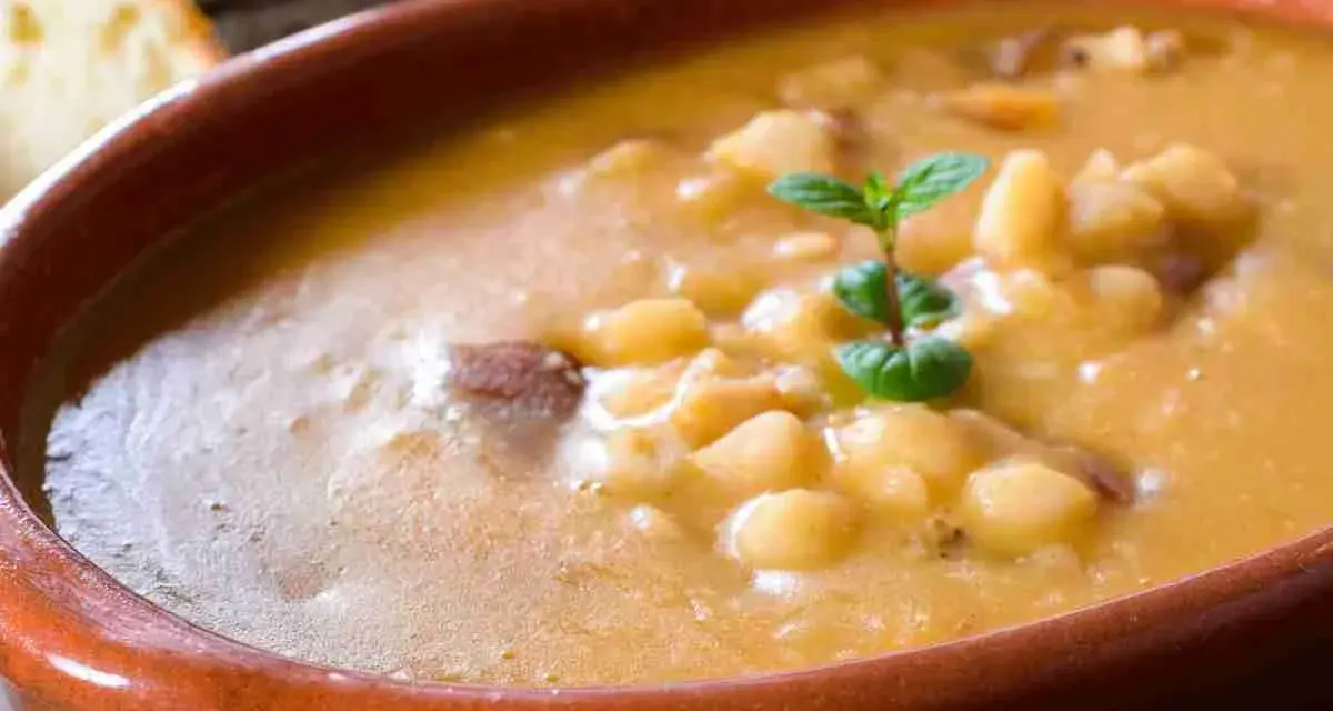 Una ciotola di terracotta piena di zuppa di fagioli cremosa, con pezzetti di carne e una fogliolina di menta. Perfetta per una cena confortante.