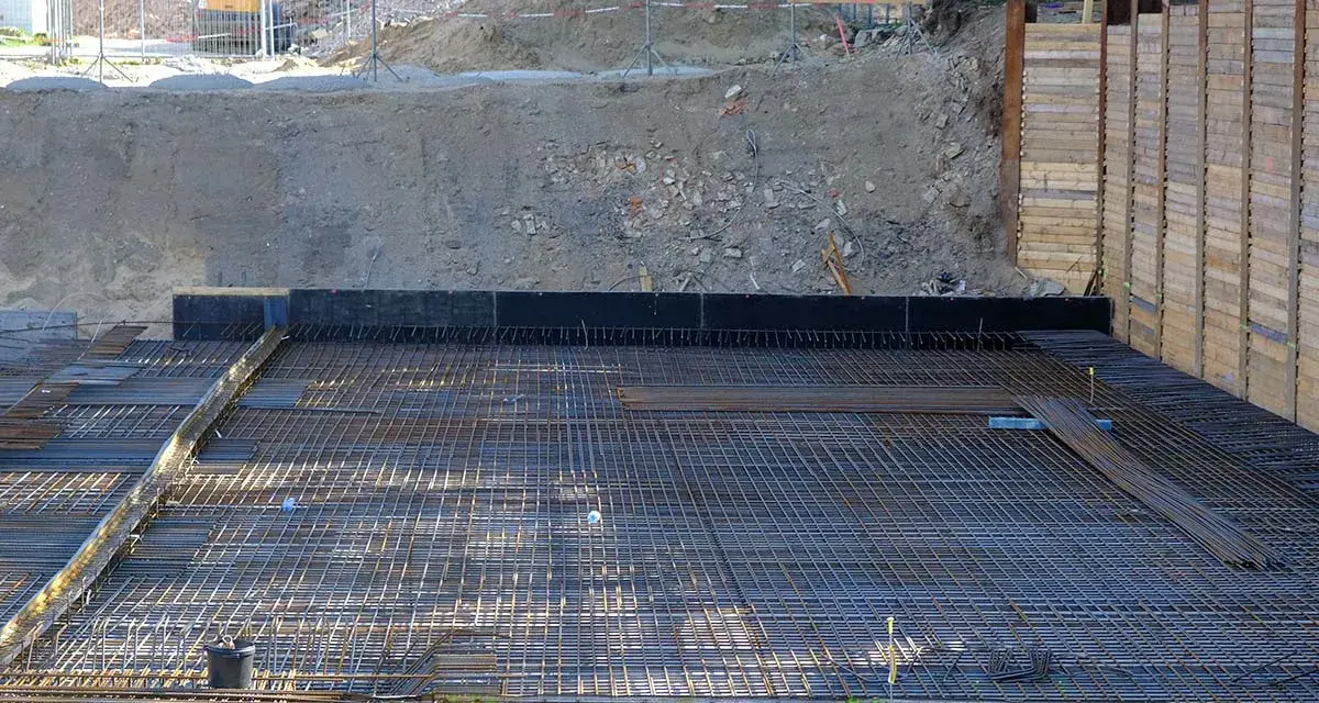 Armatura per fondazione a platea sezione. La rete metallica è pronta per il getto di calcestruzzo, con terra e muri di contenimento sullo sfondo.