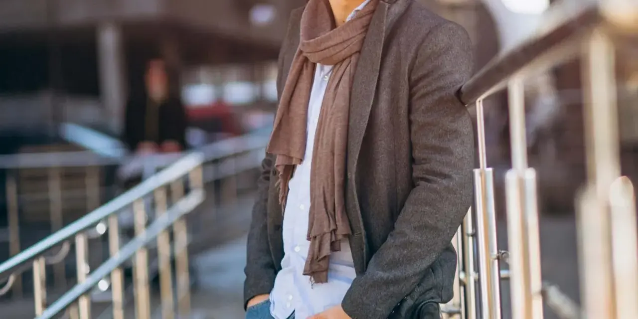 Uomo con cappotto grigio, camicia azzurra e pashmina marrone, un esempio di come indossare una pashmina in modo chic.