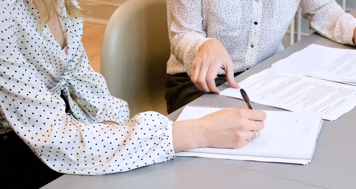 Two women in polka-dot shirts review documents. One takes notes, implying they are discussing who can see committee meeting minutes.