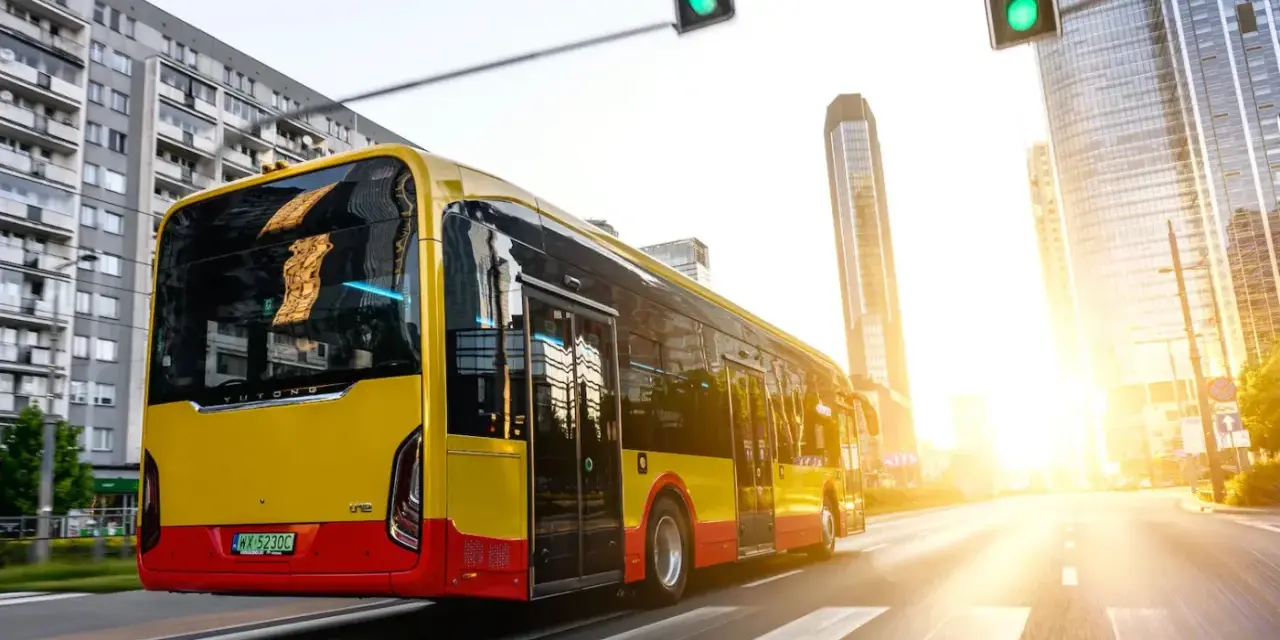 Żółto-czerwony autobus elektryczny Yutong jedzie przez miasto w blasku zachodzącego słońca.