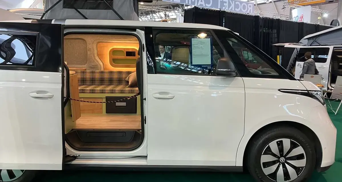 Vit ID Buzz camper van med uppfällt tak och öppen skjutdörr som visar en mysig interiör med rutig soffa och trädetaljer.