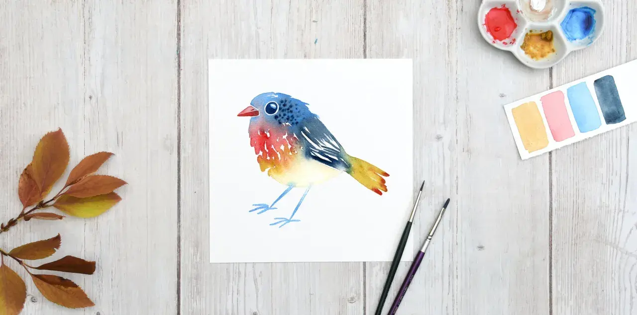 Un charmant dessin oiseau aquarelle aux couleurs vives, posé sur du papier blanc. Des feuilles d'automne et du matériel de peinture complètent la scène.