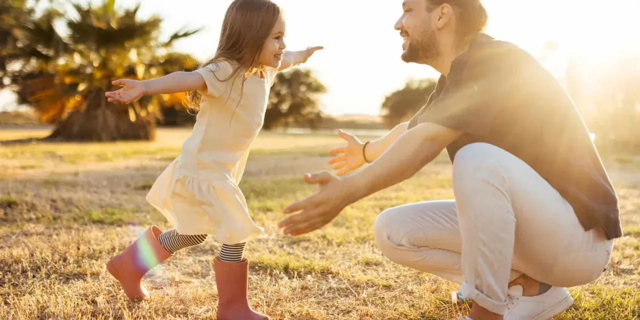 Ein kleines Mädchen rennt mit offenen Armen auf ihren lachenden Vater zu. Die warme Abendsonne beleuchtet die Szene und unterstreicht die liebevolle vater tochter beziehung.