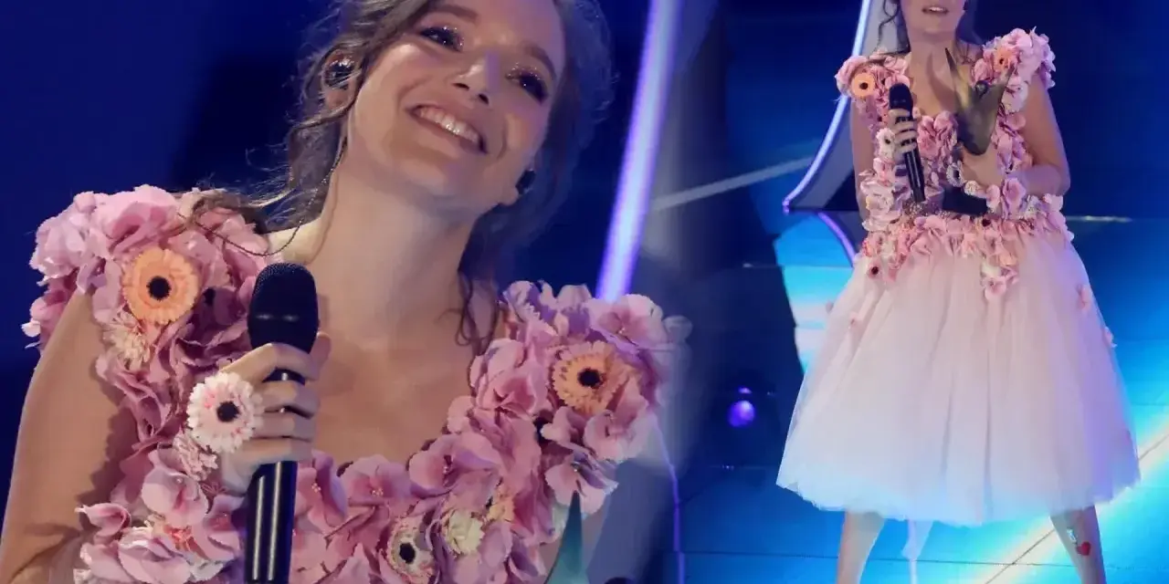 Zuzanna Irena Jurczak w kwiecistej sukni na scenie, trzyma mikrofon i uśmiecha się do publiczności.