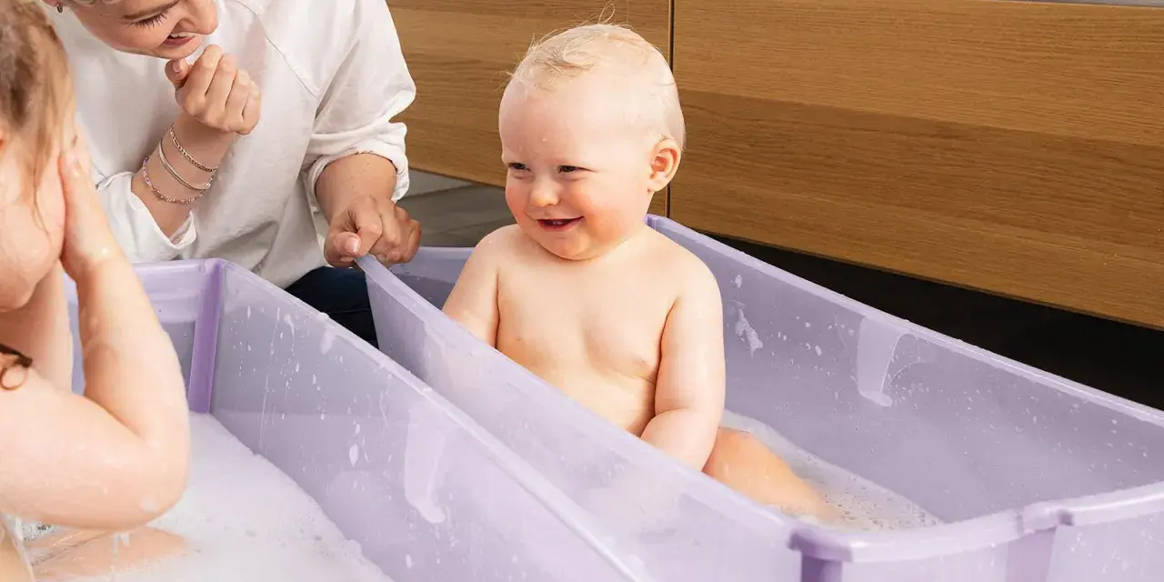 Uśmiechnięty bobas w fioletowym krzesełku do kąpieli, mama patrzy z czułością.