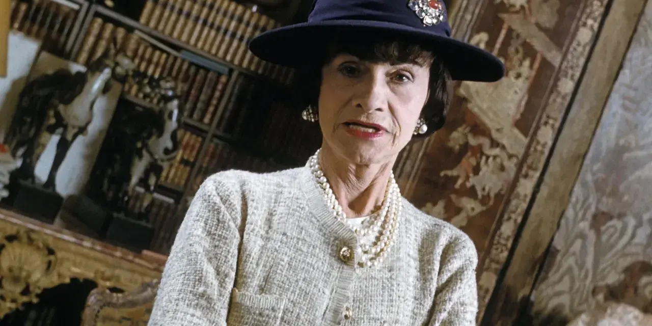 Iconica stilista Coco Chanel, con cappello blu e perle, in un ambiente elegante con libri e decorazioni.