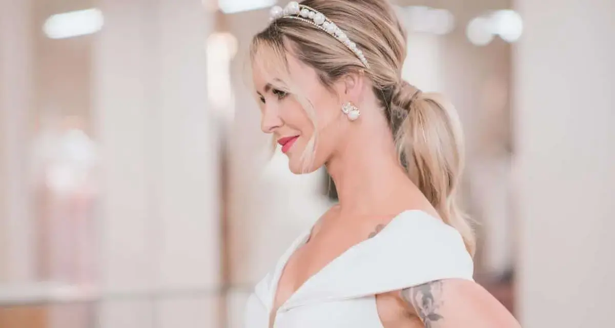 Elegancki kucyk ślubny z perłową opaską i kolczykami. Panna młoda w białej sukni z dekoltem.