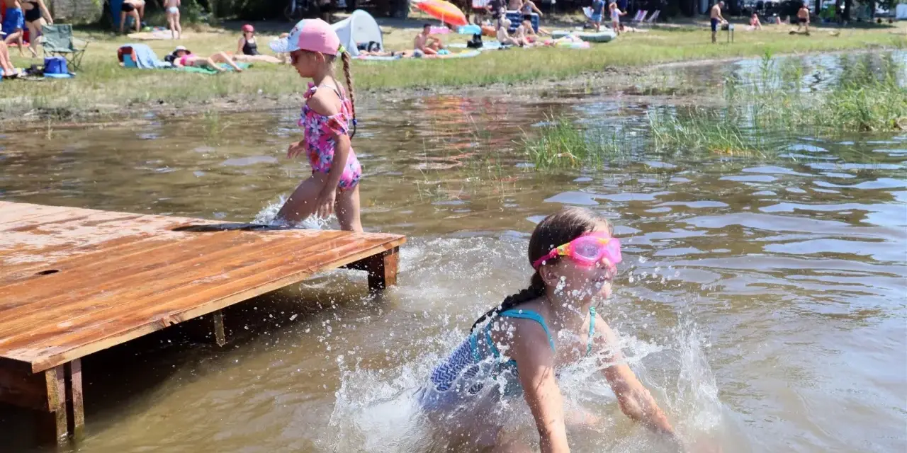 Dwie dziewczynki pluskają się w wodzie przy drewnianym pomoście. W tle plażowicze i drzewa. Wakacje nad jeziora lubelskie.