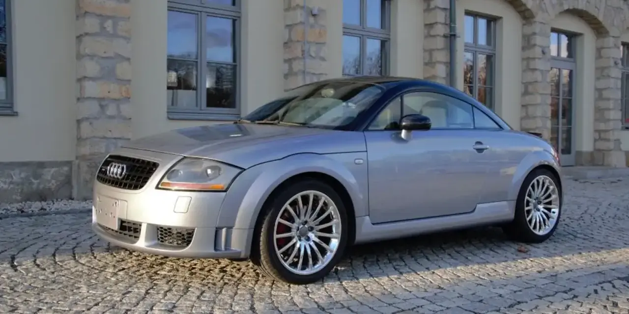 Silberner Audi TT 2005 mit schwarzem Dach parkt auf Kopfsteinpflaster vor einem Gebäude mit Steinbögen.