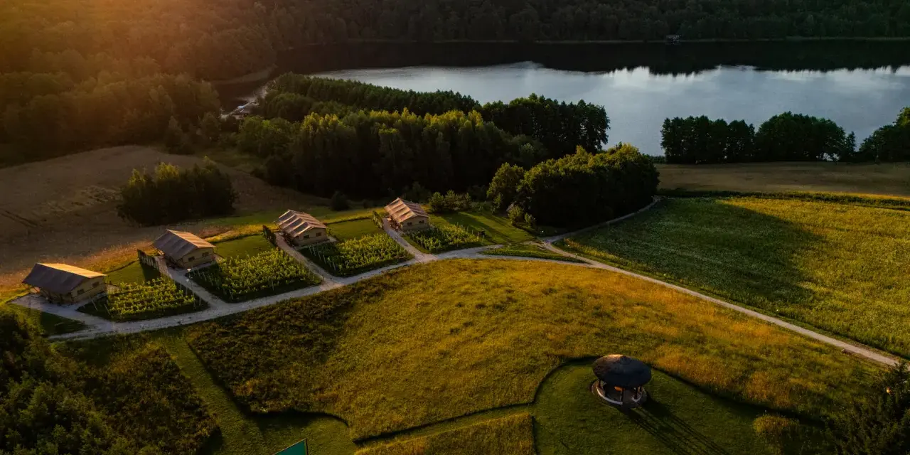 Idealne miejsce, gdzie pojechać na weekend: urokliwe domki nad jeziorem, otoczone zielenią i polami, w złotym świetle zachodzącego słońca.