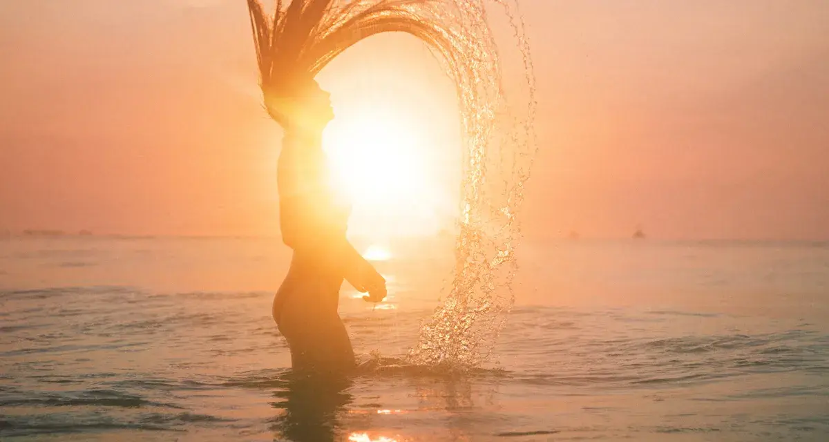 Silhouette d'une femme jetant ses cheveux dans l'eau au coucher du soleil, avec un effet de lens flare photoshop.