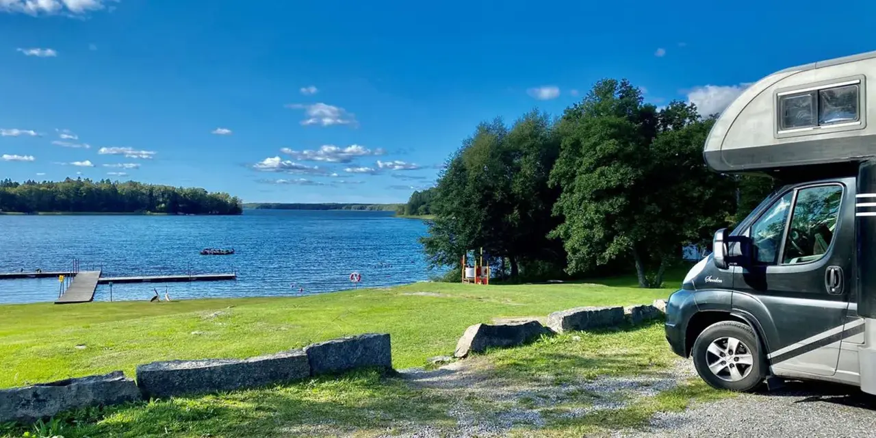 Husbil vid en sjö med brygga, perfekt för fricamping Sverige. Blå himmel och grönska.