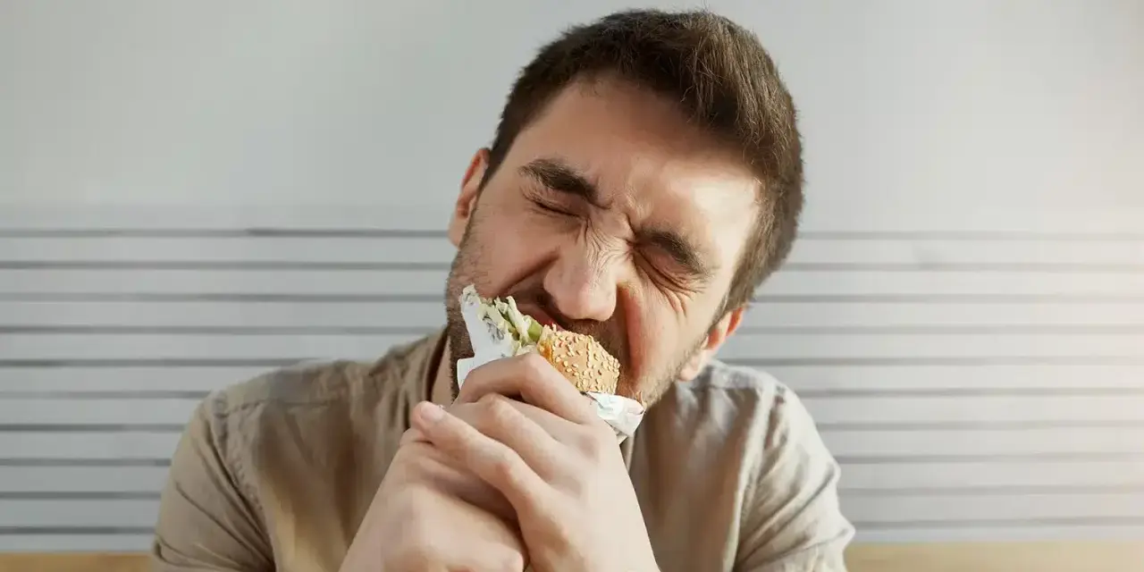 Uomo con espressione sofferente mangia un panino, forse per alleviare lo stress, ma teme di ingrassare.