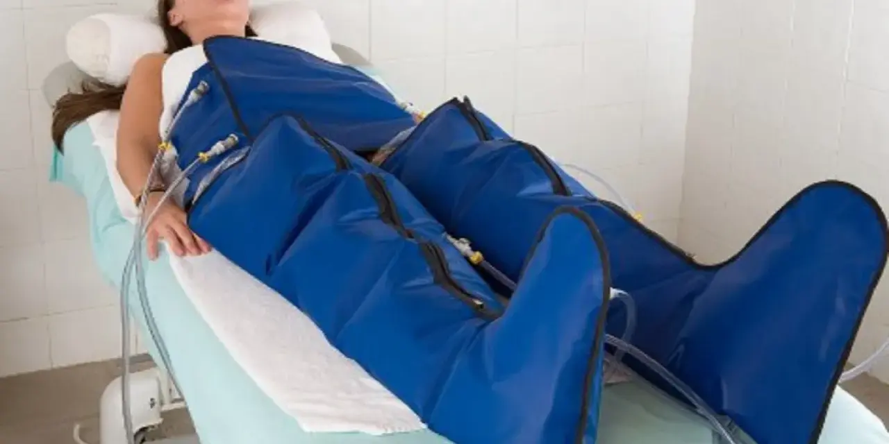 Kobieta leży na łóżku, jej nogi są owinięte w niebieskie mankiety urządzenia do mechanicznego drenażu limfatycznego.