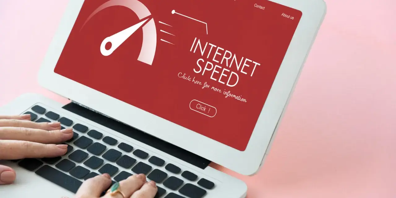 Schermo di un laptop con indicatore di velocità internet su sfondo rosso. Le mani digitano sulla tastiera, frustrati dall'internet lento.