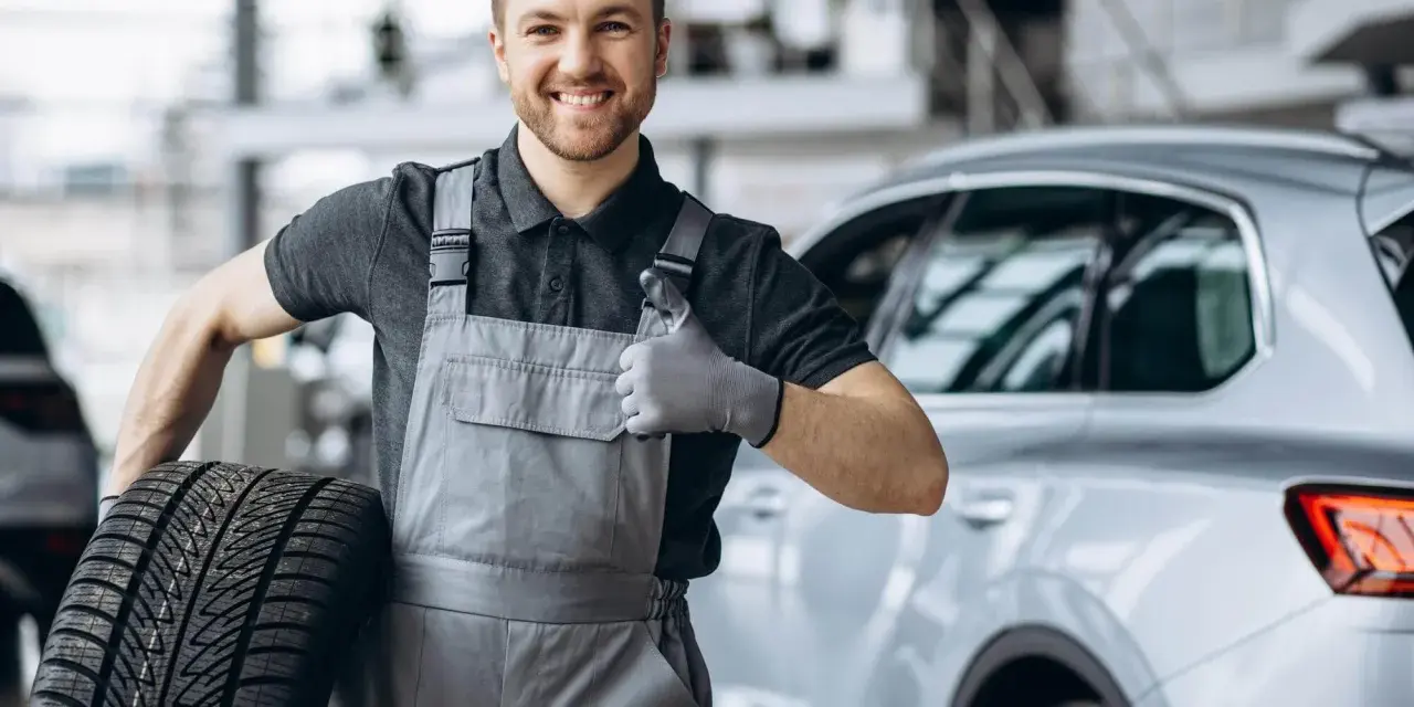 Meccanico sorridente con una gomma primo equipaggiamento in mano, fa il pollice in su accanto a un'auto.