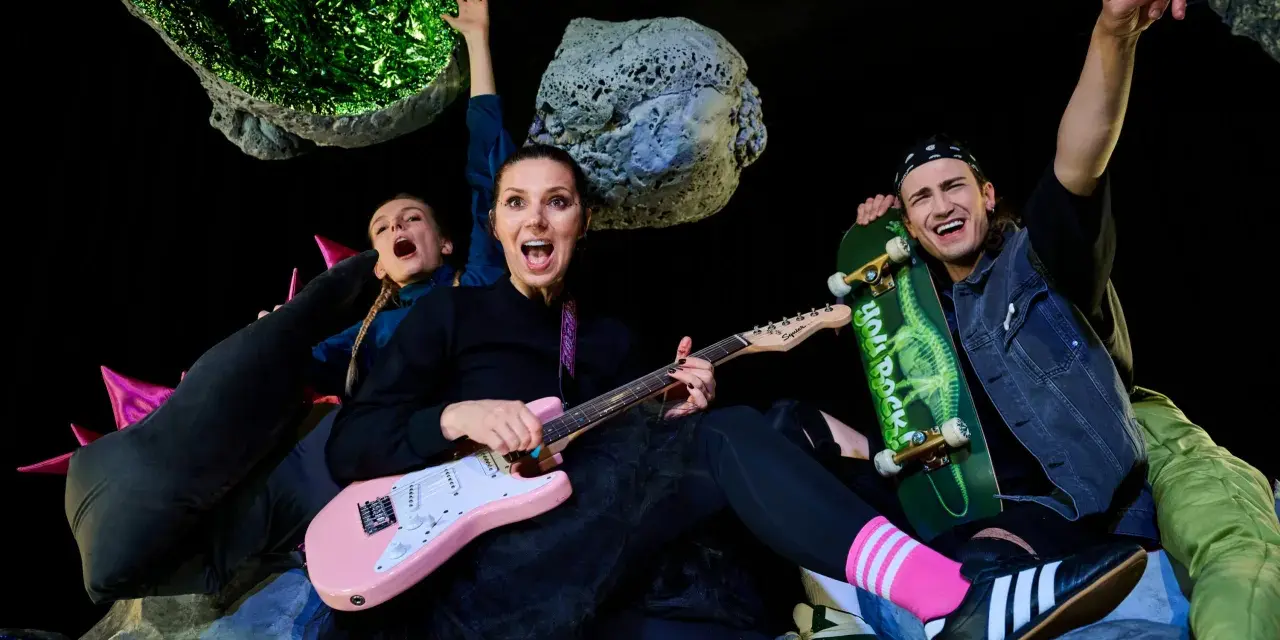 Energiczna ekipa w kosmicznej scenografii, grająca polski folk rock. Kobieta z różową gitarą, mężczyzna ze skateboardem, wesołe miny i podniesione ręce.