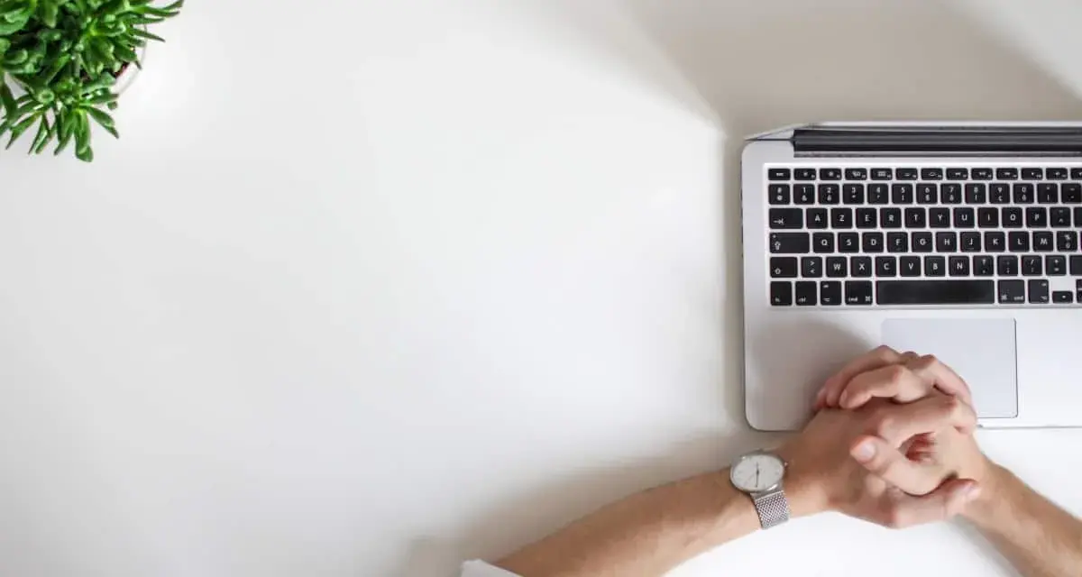 Minimalistyczne biurko z laptopem, zieloną rośliną i dłońmi złożonymi w geście oczekiwania. Idealne do wizualizacji pracy kreatywnej i renderowania.