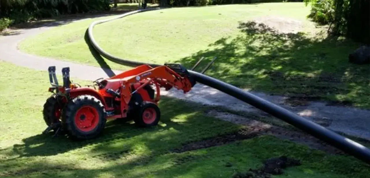 A tractor with a front loader is laying a long, flexible HDPE pipe across a grassy area.