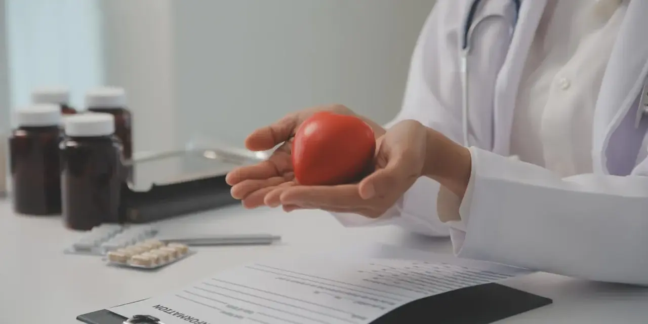 Lekarz trzyma w dłoniach czerwone serce, symbolizujące zdrowie. Na stole leżą leki i dokumentacja medyczna, sugerując rozmowę o tym, po jakim czasie spada cholesterol.